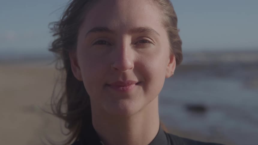 Closeup portrait of a woman at the beach softly lit with a gentle smile wind teases a few strands of hair and the ocean is blurred in the background under a clear sky