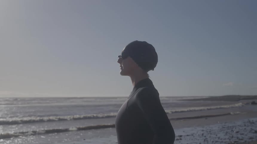 The swimmer is standing on the sand looking out over the water