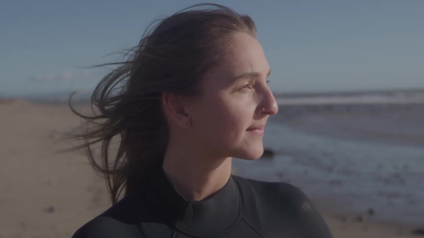 A medium close side view shows a young woman with long dark hair blowing in the wind wearing a black wetsuit and smiling as she looks out toward the sandy beach and ocean
