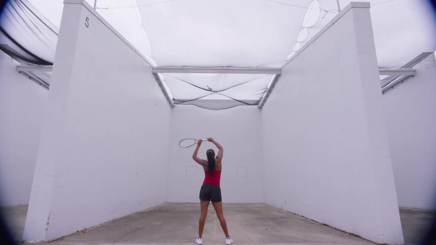 Woman practicing tennis alone on a court surrounded by high walls