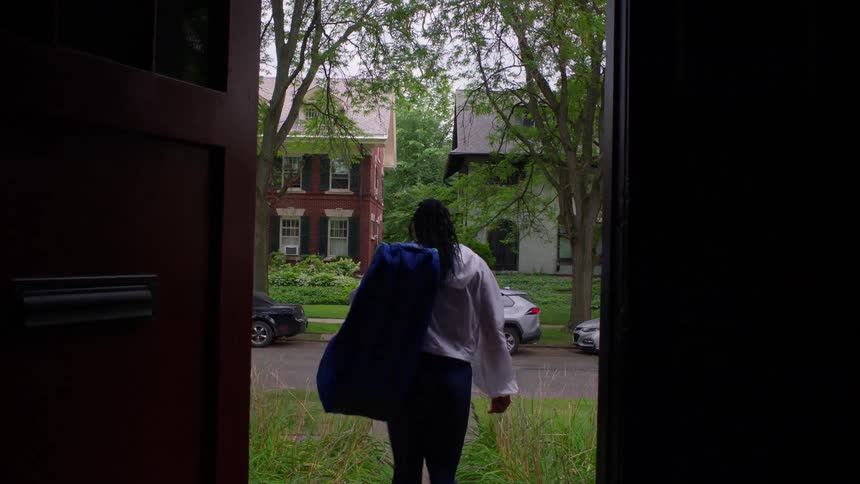 A young woman carrying a backpack walks out of a building into the street at midday where cars are parked along the roadside