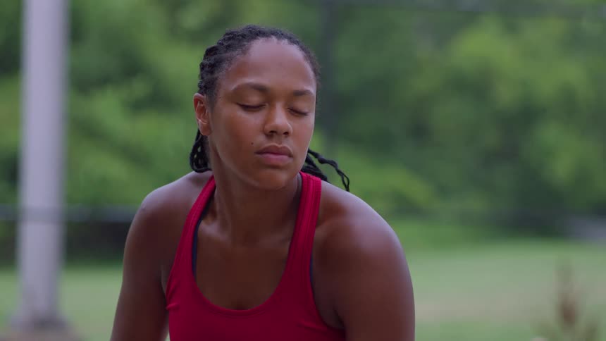 A woman with braided hair wearing a red top who transitions from a moment of closedeye rest to opening her eyes and raising a tennis racquet above her head in a continuous motion set outdoors against a green and white background