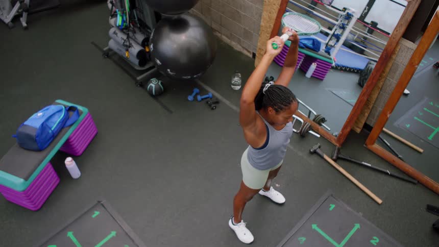 A woman standing on a fitness facility raising a racquet above her head surrounded by various pieces of gym equipment