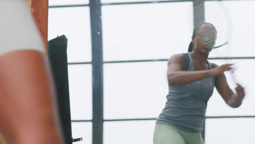 The reflection of a young woman with long braids wearing a gray tank top and pale green shorts is seen in focus as she repeatedly swings a tennis racket in a bright gym with large grid windows while her actual body is visible in the foreground