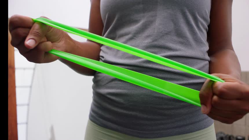 A woman hold a green rubber exercise band and repeatedly stretch and relax the band in front of the body on an indoor setting