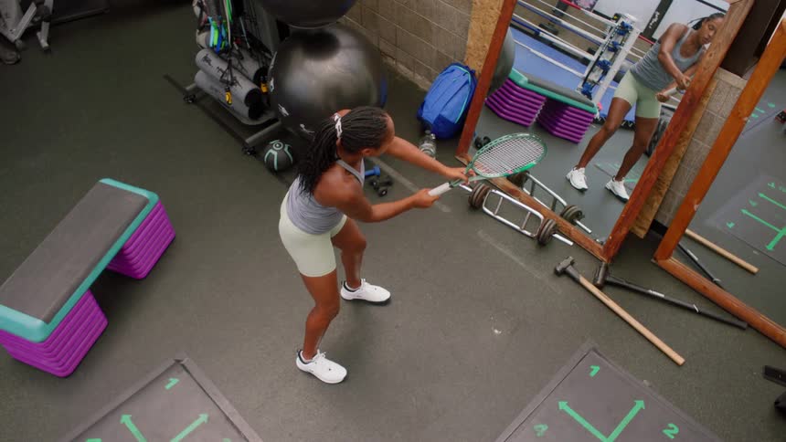 A woman practices tennis in front of a mirror in a gym