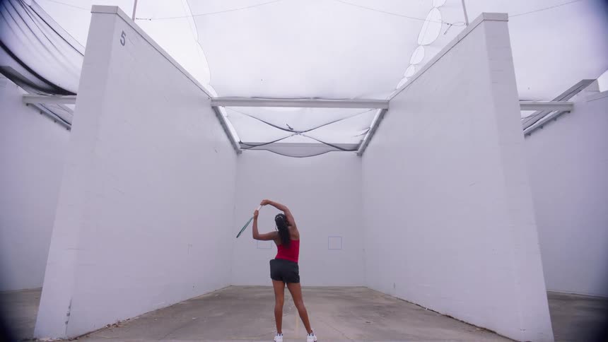 Woman practicing tennis swings alone on a court surrounded by walls