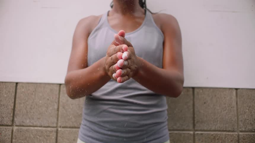 A person with bare arms and a tank top standing against a tiled wall extending their hands forward and clapping them together once