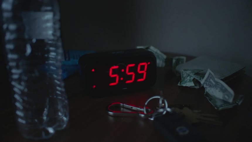 A digital alarm clock with bright red numerals reading 600 sits on a dark bedside table next to a plastic water bottle keys and crumpled dollar bills hit by a hand
