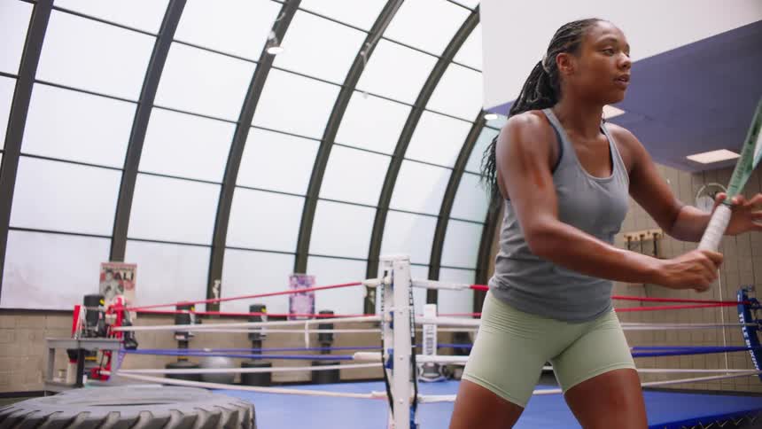 A young woman stands next to a large tire inside a gymnasium with a boxing ring and repeatedly swings a tennis racquet in an overhead motion