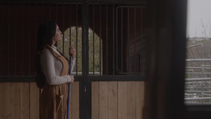 A woman in a tan dress and longsleeve top stands inside a horse stall holding a stick and looking toward the open doorway soft overcast light falls across the wood panels and metal bars