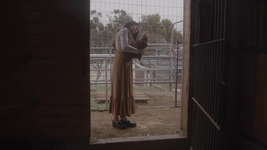 Woman holds a chicken in a barn doorway by a fenced pen