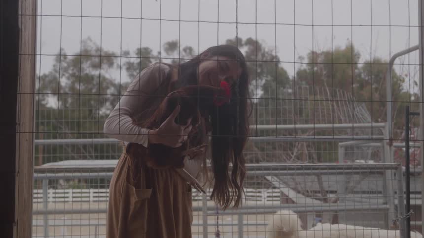 Woman smiles while holding a rooster in an outdoor enclosure