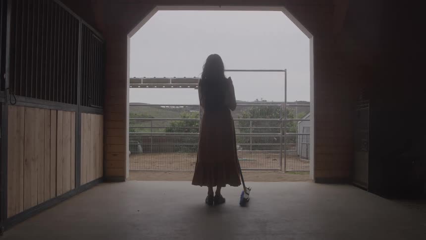 Woman stands with a broom while looking at the landscape outside the barn