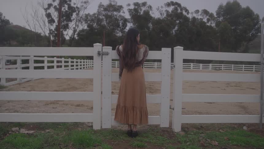 A woman in a long tan dress stands at a white paddock fence her hands on the gate