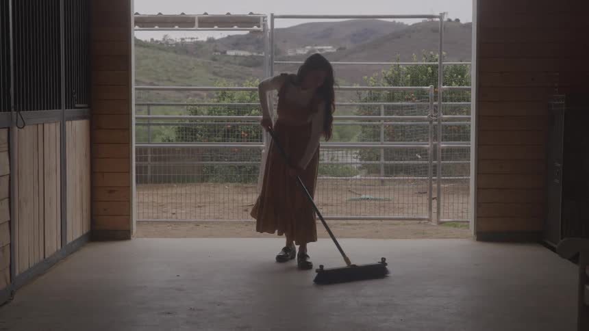 Woman sweeps the barn floor with hills in the background