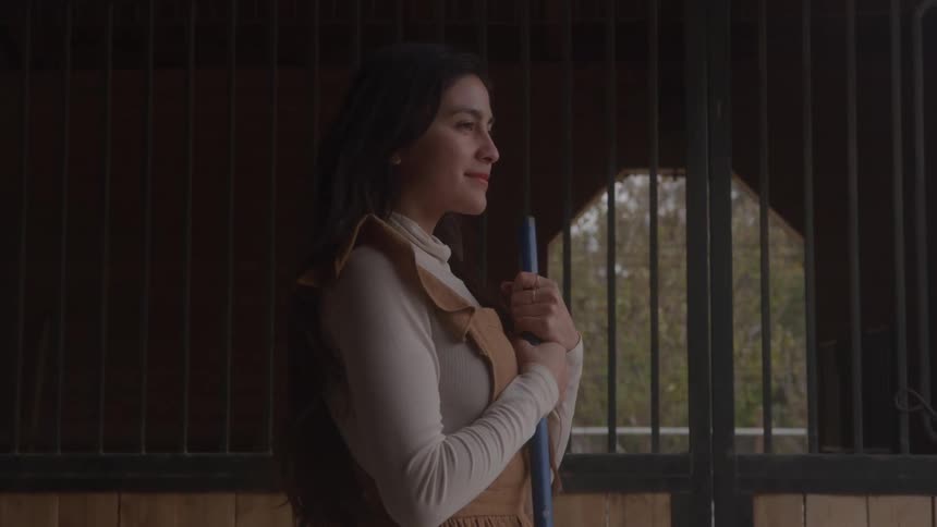A young woman with long dark hair wears a white longsleeve shirt and a tan jumper smiling while leaning on a blue broom handle inside a dark stable stall facing the sunlit outdoor window