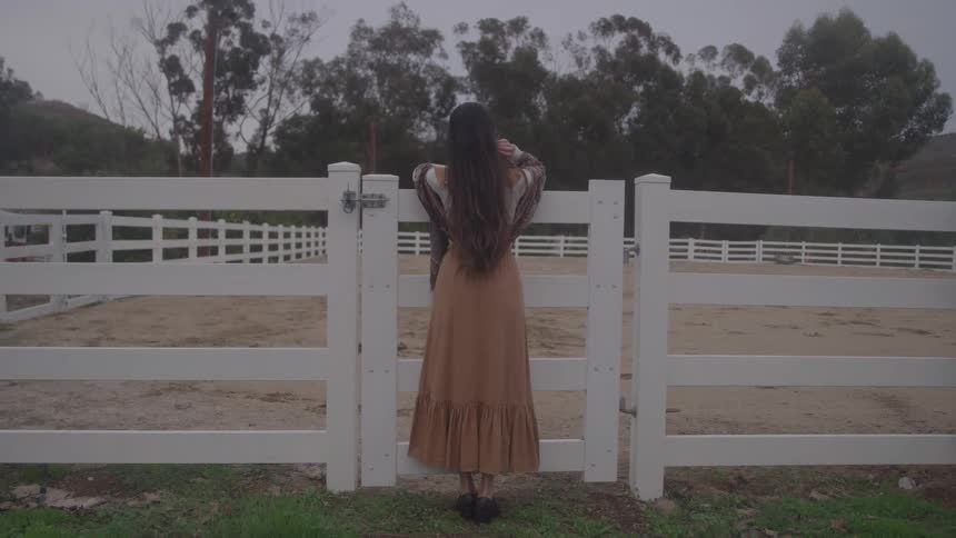 A woman with long dark hair wearing a long dress standing with her back to the camera and leaning against a white wooden fence that encloses a dirt field with trees covering the background hillside