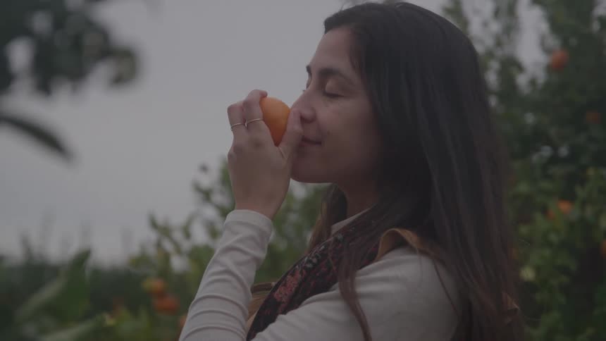 A woman with long dark hair holds an orange to her nose inhaling the scent with her eyes closed in an orchard