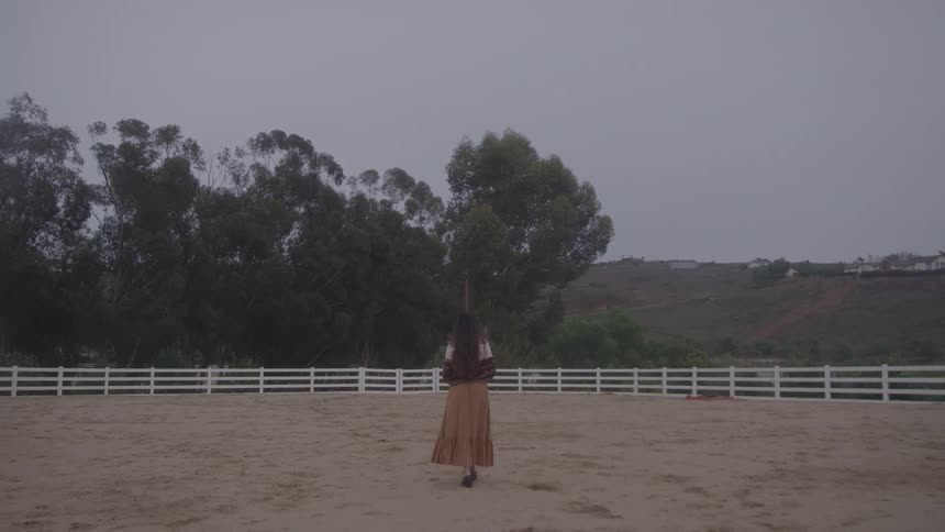 Woman walks across a dirt arena toward a fence on an overcast day