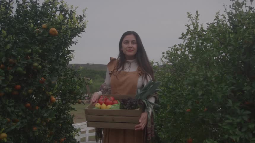 A smiling woman in an apron stands in an orchard holding a crate overflowing with freshly harvested vegetables and fruit