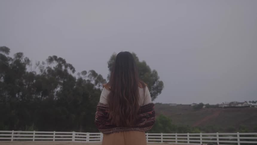 A person with long hair standing motionless in a fencedin dirt area looking out over a hillside covered in trees and distant houses beneath a gray clouded sky