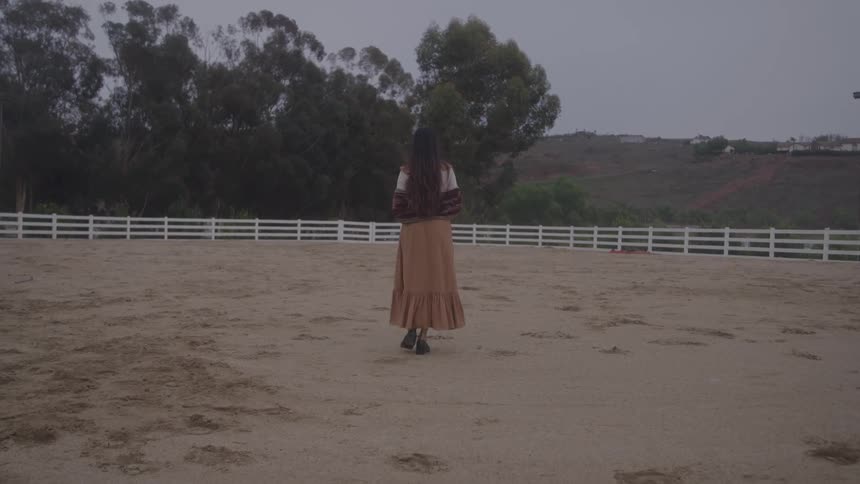 A woman walks across a dirt arena toward the trees and hills
