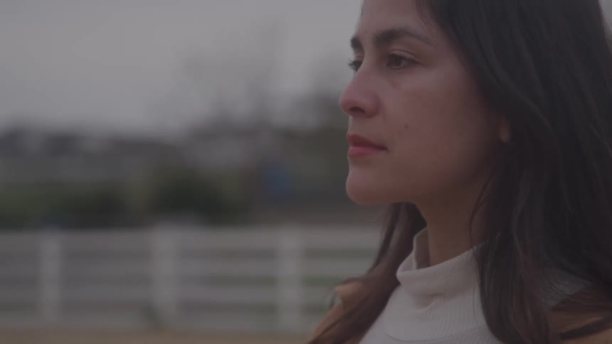 A woman with long dark hair wearing a white shirt looking to the left with a neutral expression positioned outdoors against a blurred background containing a horizontal white fence