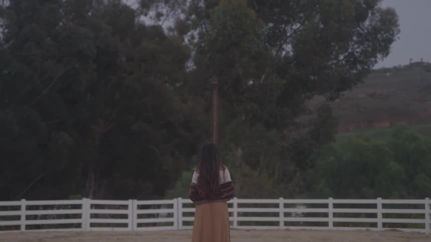Woman stands on a ranch overlooking trees and hills at sunset