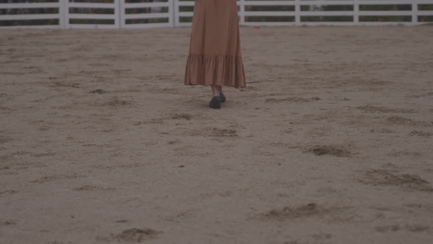 Mediumclose view of the lower half of a person wearing a long skirt and dark shoes walking away from the camera across a dirtcovered surface toward a white slatted fence