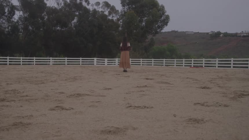 A young woman with long dark hair wears a tan dress and a white shawl as she stands in the center of an empty dirt horse corral enclosed by a white wooden fence on a cloudy day