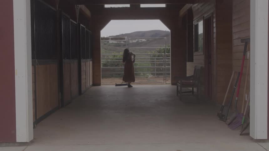 Woman sweeps barn floor with hills and orange trees outside