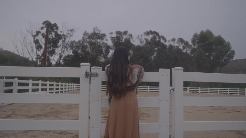 A woman in a long tan dress stands at a white paddock gate with her back turned calm and contemplative