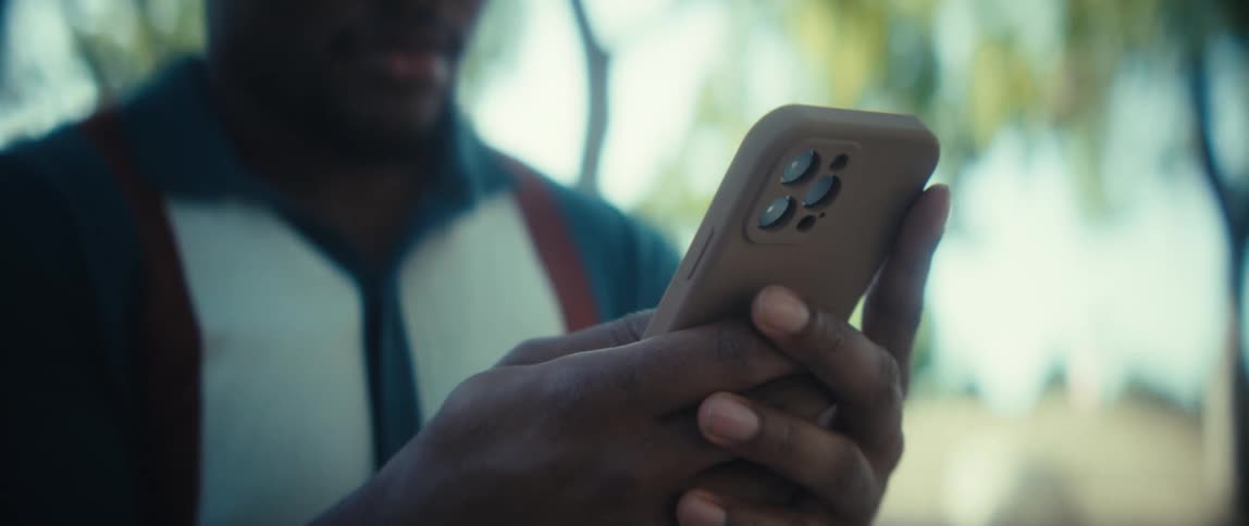 A close view shows a man wearing a teal polo shirt holding a smartphone with a brown protective case and scrolling while standing outdoors with green foliage in the blurred background