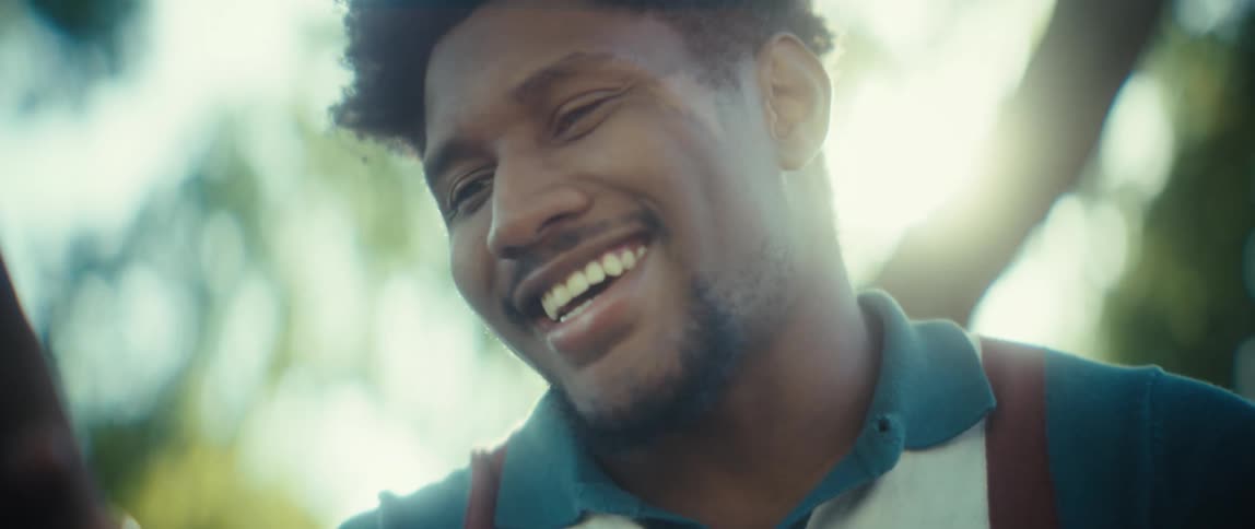 A young man with short curly hair smiles and laughs brightly into the distance in a sunlit outdoor setting