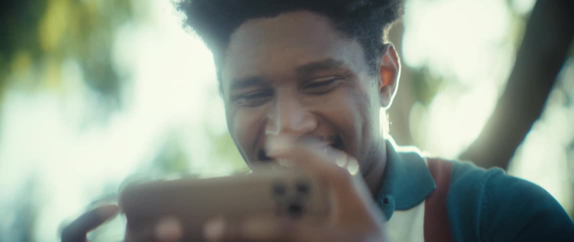 A young man holds a mobile phone close to his face and smiles while viewing content outdoors with bright light streaming from behind him