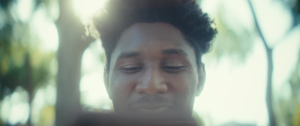 A close view of a young man with dark curly hair talking on a smartphone smiling and laughing outside in a natural setting with bright sunlight filtering through the green trees in the softfocus background
