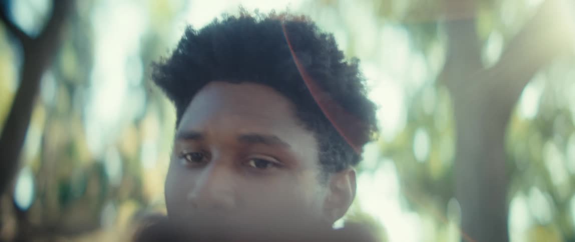 A young man sits under a tree in a public park at midday holding his phone in his hands while engaged in a video conversation