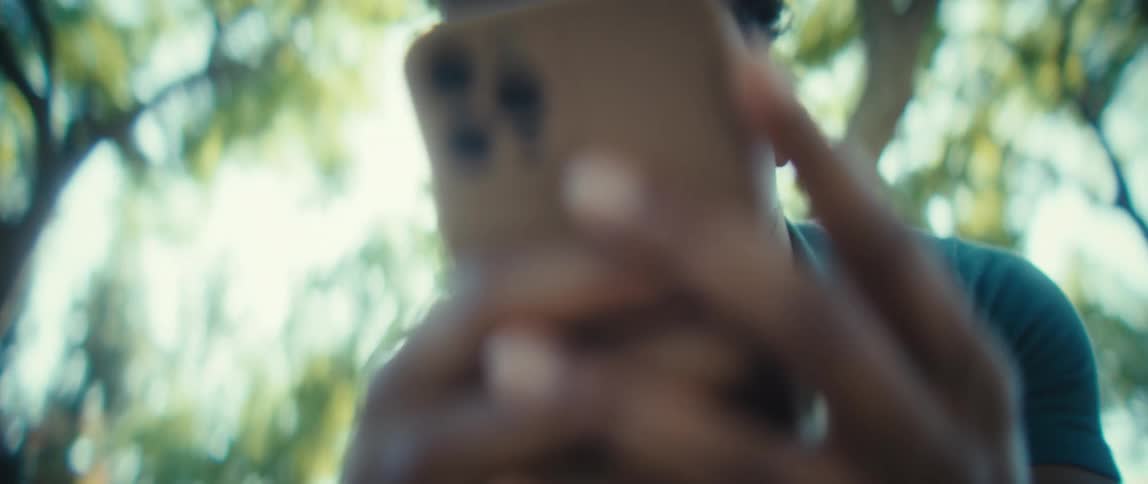 A closeup view shows a young man with short dark hair wearing a collared tshirt using a smartphone outdoors smiling while interacting with the device against a sundappled green background