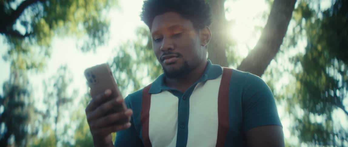 Man holds a smartphone while looking intently at the screen under sunlight filtering through the trees