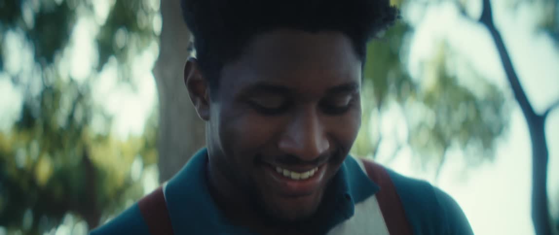 A black male with a collared shirt and suspenders visible in soft daylight is using a phone while smiling