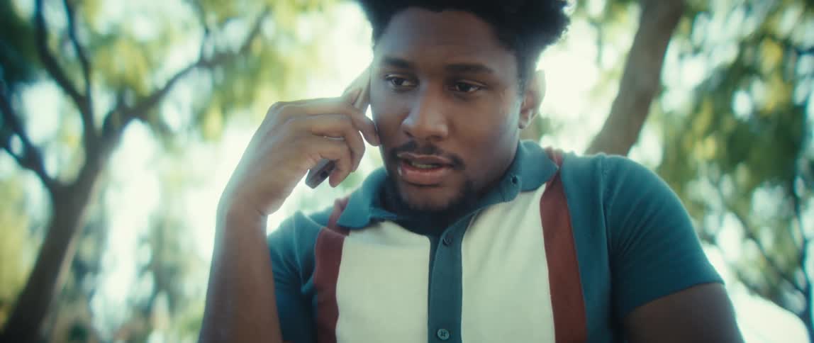 A young black man with dark curly hair wearing a teal and white polo shirt holds a smartphone to his ear and speaks while standing outdoors in the bright sunlit shade of a tree