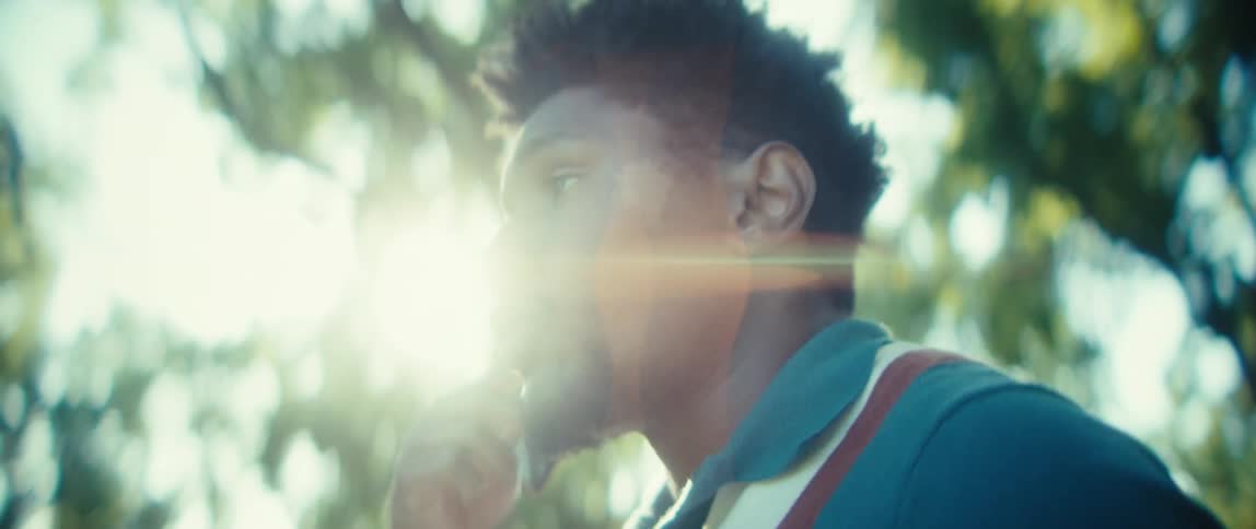 A close side view shows a young black man with short dark hair wearing a teal polo shirt talking on a smartphone outdoors with bright sunlight filtering through the green trees in the background