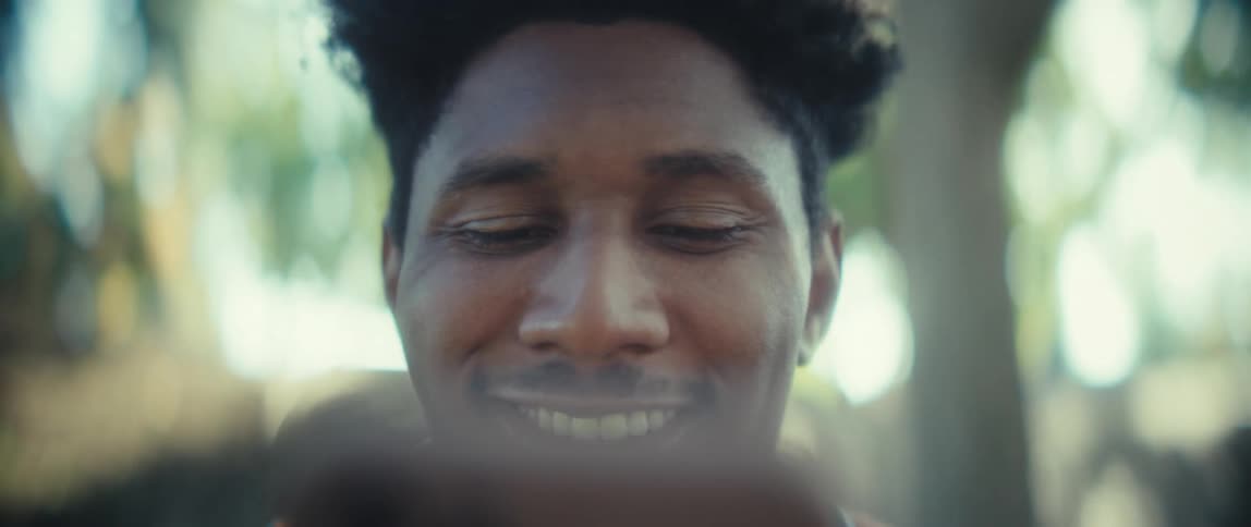 A young man looking to a smartphone and laughing in an outdoor setting with trees in a blurred background