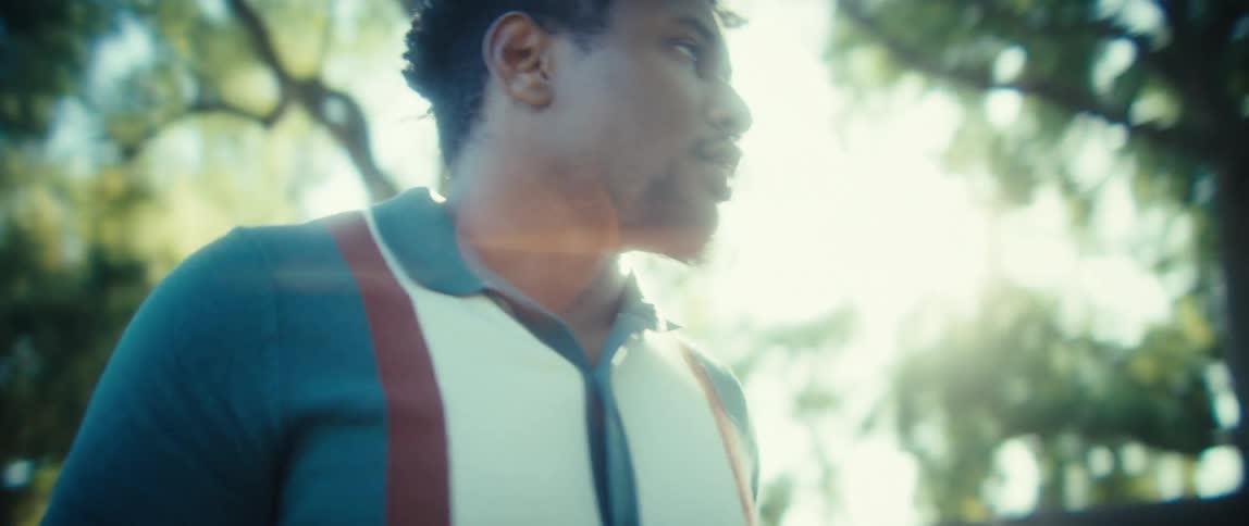 A man wearing a teal and white polo shirt with suspenders looks at and uses a smartphone before turning and walking away from the camera into a sunlit park with green trees