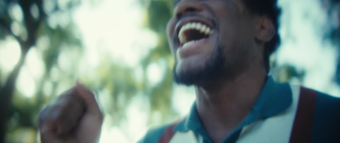 A man with a short beard holding a smartphone with a wideopen mouth expressing intense positive emotion against an outoffocus backdrop of green trees