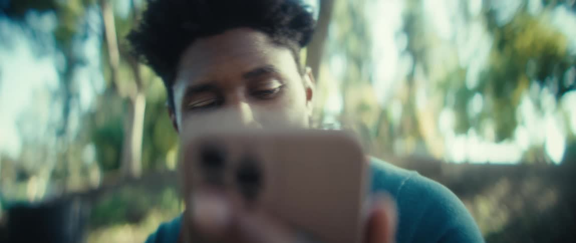 A man with dark short hair holding a cell phone close to his face with his expression changing from neutral to a wide smile