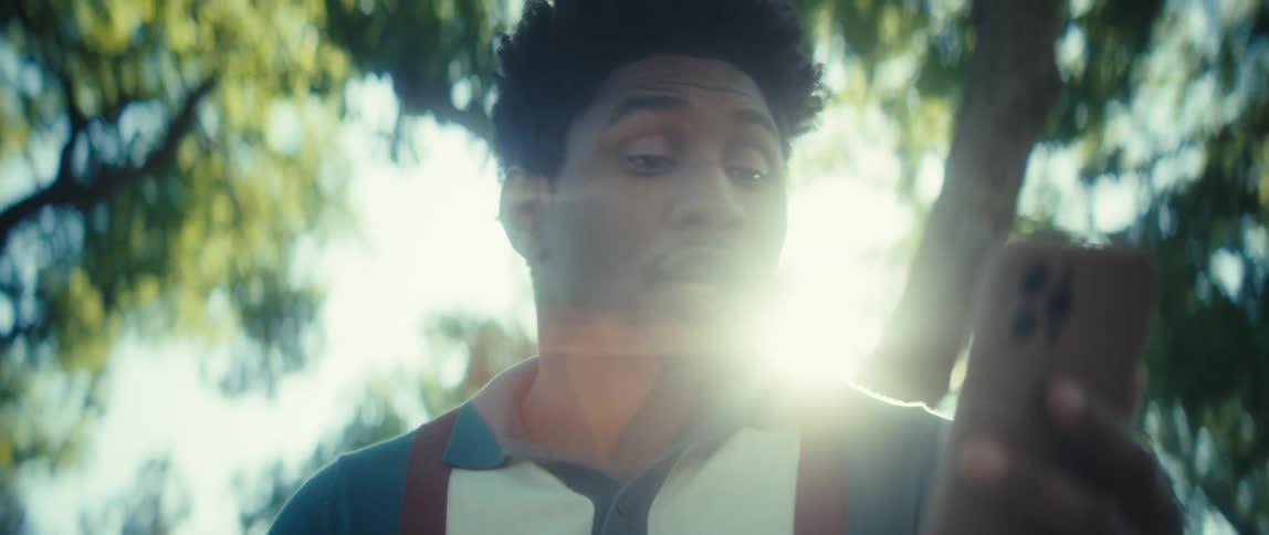 A man holding a smartphone and reacting to something on the screen with a bright sun flare dominating the right side against a blurred backdrop of green trees