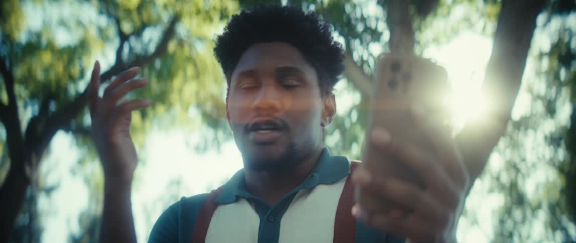 A young man having a conversation looking to a smartphone in an outdoor setting with trees in the background