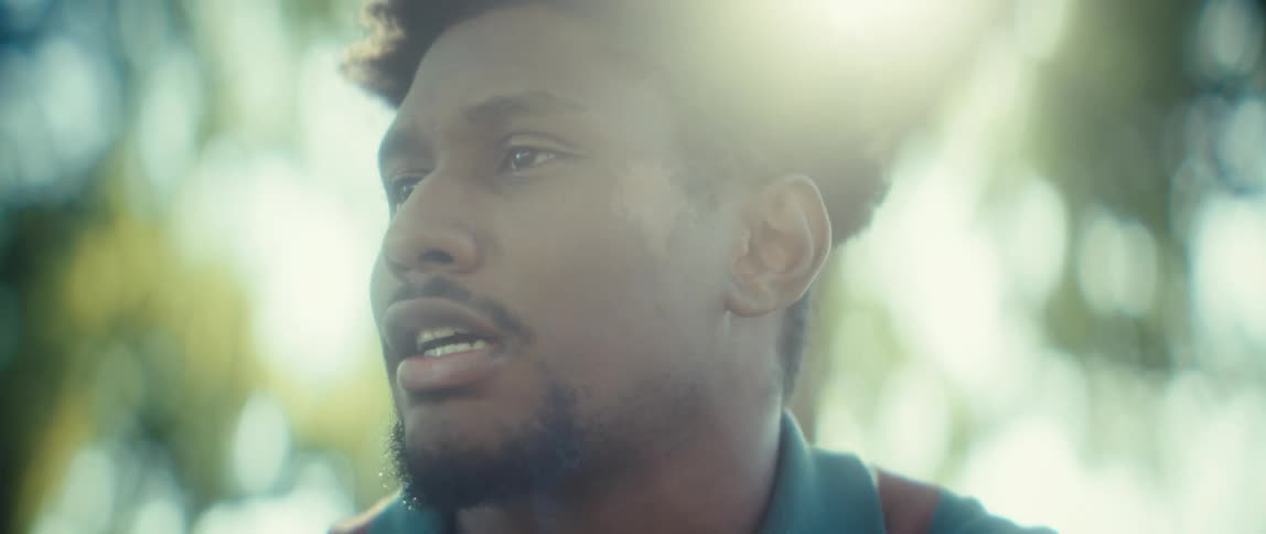 A close view shows a black man wearing a teal polo shirt talking while holding a smartphone to the side of his face in bright outdoor light with blurred green foliage in the background